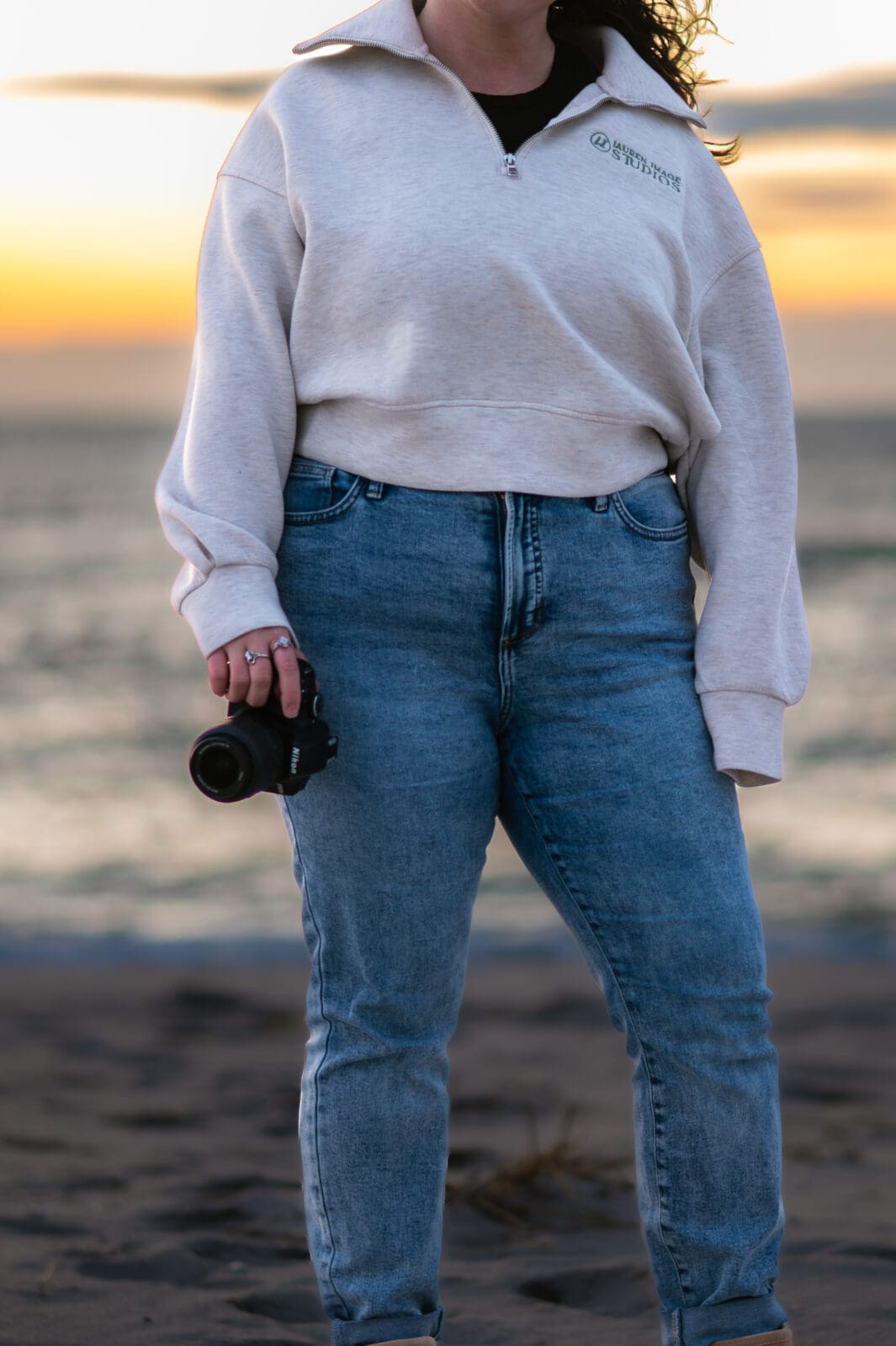 A close up of a photographer holding her camera on the beach wearing a sweatshirt with her logo for Lauren Image Studios.