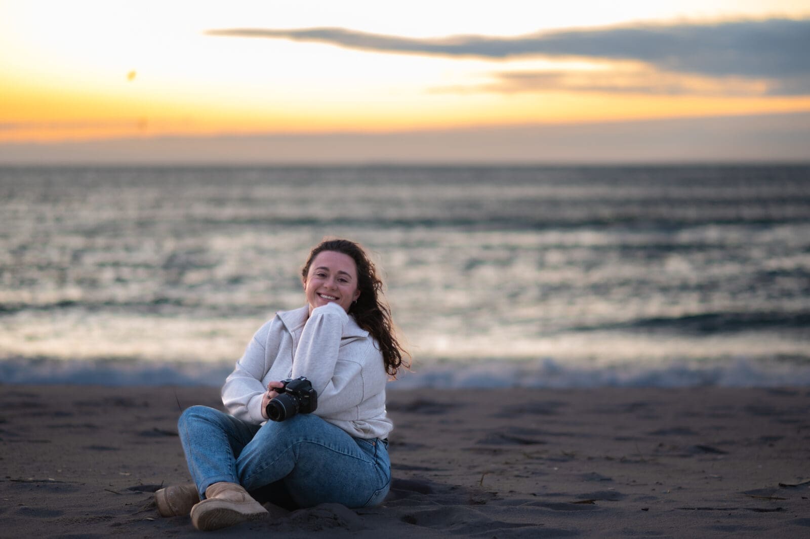 A photo of a photographer sitting on the beach with her camera in her hand at sunrise.