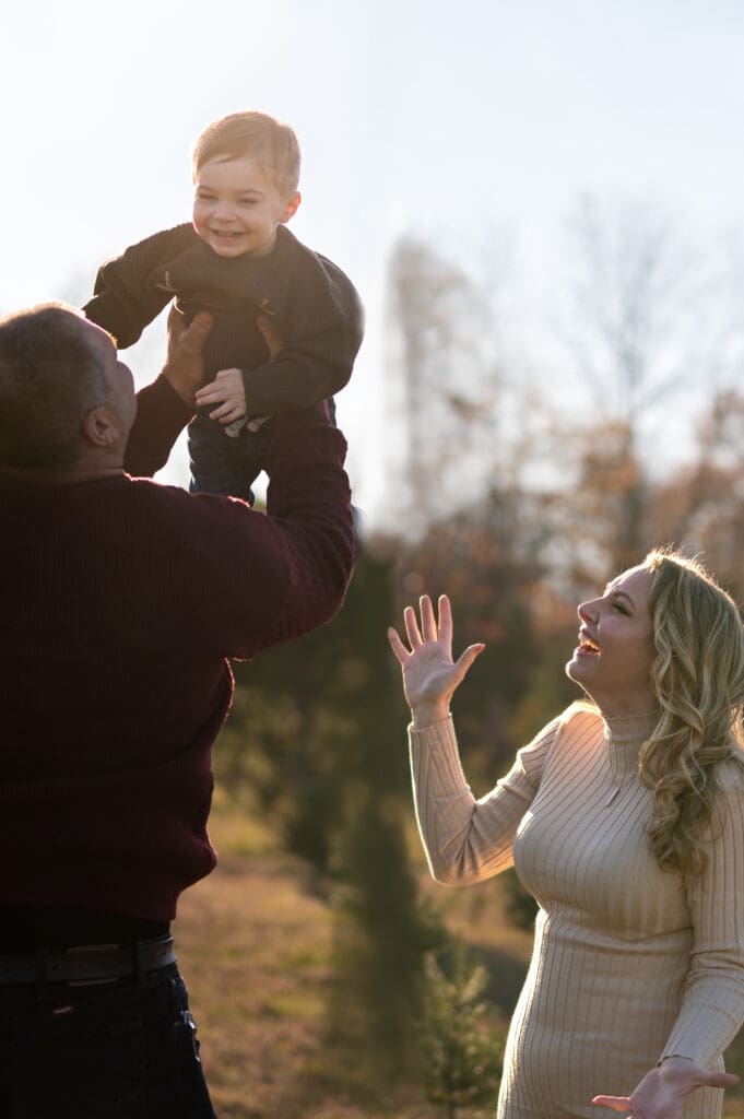 A dad throws his son in the air while a mom is looking on next to them at a Christmas tree farm in Sussex County NJ