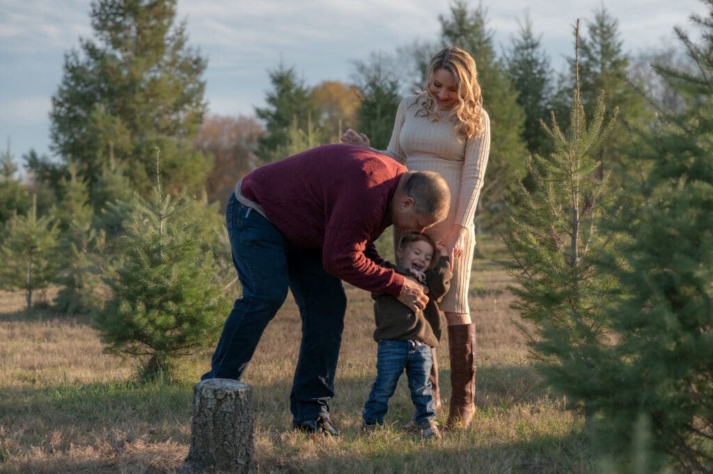 A dad bends down to tickle his laughing son while a mom holds her son's hand at a Christmas tree farm in Sussex County NJ
