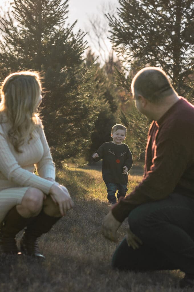 Two parents squat in the front of the frame looking back at their toddler who is running toward them at a Christmas tree farm in Sussex County NJ