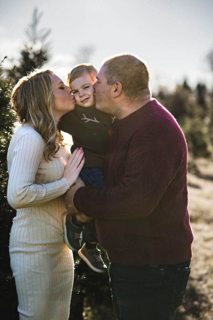 Two parents hold their toddler between them, each parent giving their son a kiss on the cheek at a Christmas tree farm in Sussex County NJ