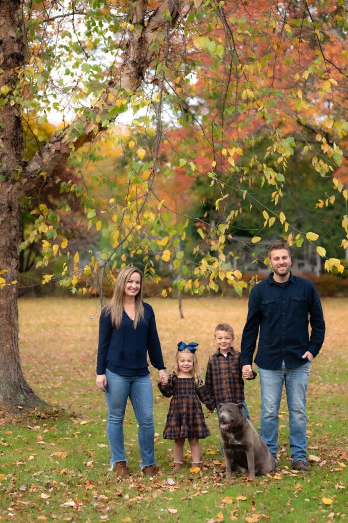 A family of four with their dog in a park standing under a tree in the fall.