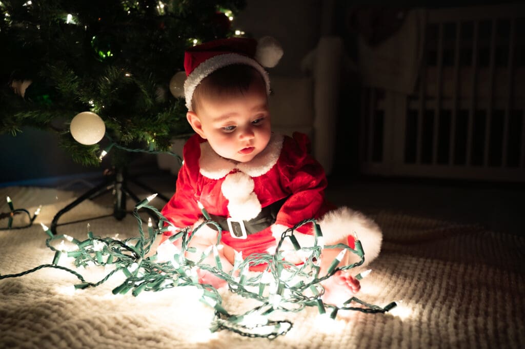 A baby dressed as Santa Claus sitting underneath the Christmas tree with Christmas lights around her.