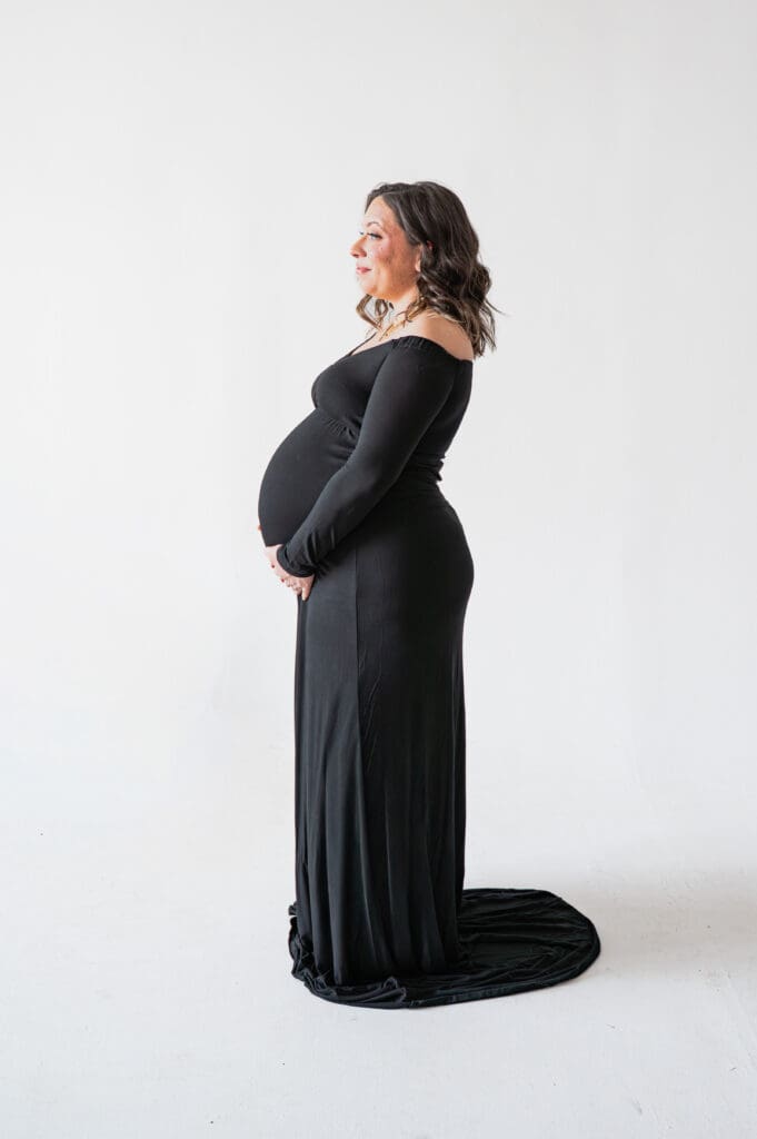 A pregnant woman stands facing the light of an open window in a studio with a white backdrop.