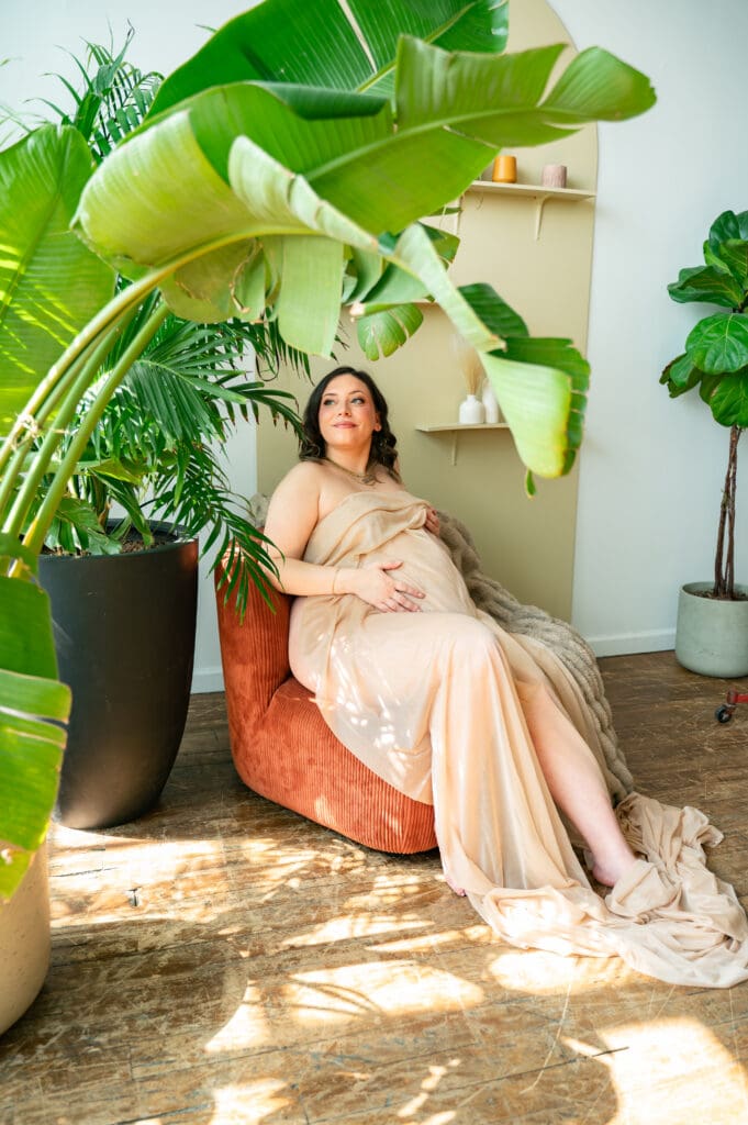 A pregnant woman is draped by a nude sheet and framed by green plants as she sits in an orange chair looking off in the distance behind the camera.