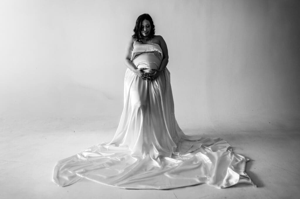 A black and white photo of a pregnant woman sitting on a stool with a satin sheet draped over her body.
