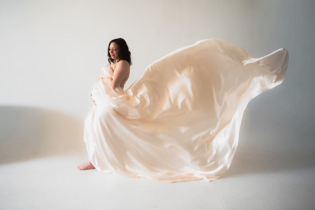 A pregnant woman is standing profile to the camera with a satin sheet flowing in the wind behind her.