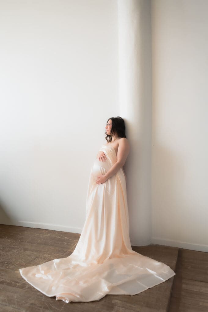 A pregnant woman covered by a nude satin sheet is leaning against a wall, looking away from the camera toward the natural light of the window.