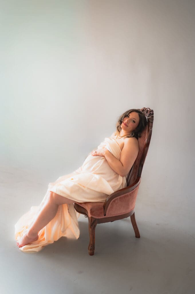 A pregnant woman sits in a chair with a satin sheet covering her, one leg is showing, and she has her head tilted back on the chair looking directly at the camera.