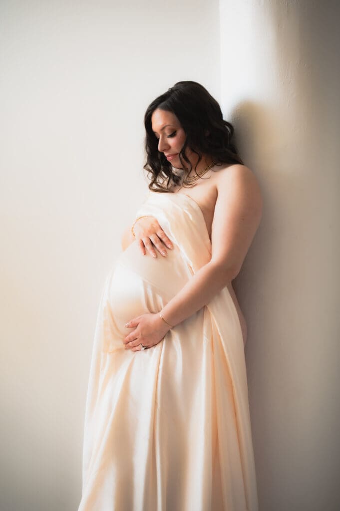 A pregnant woman leans against a white pole holding her belly covered by a nude satin sheet. She is looking down at her belly. 