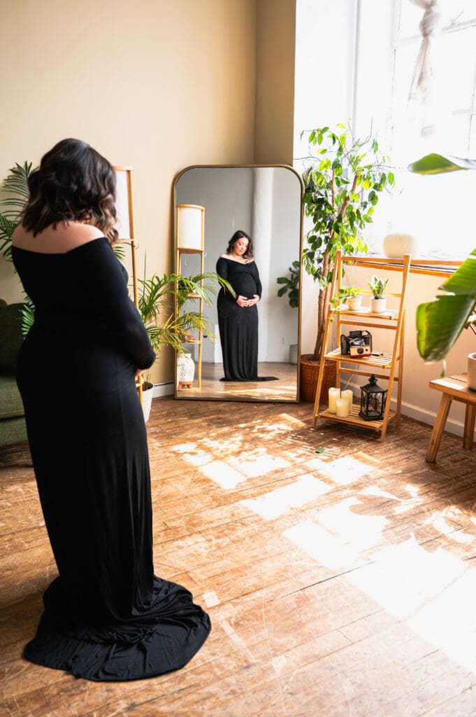 A pregnant woman in a black, floor-length dress, standing in front of a mirror, admiring her growing belly.