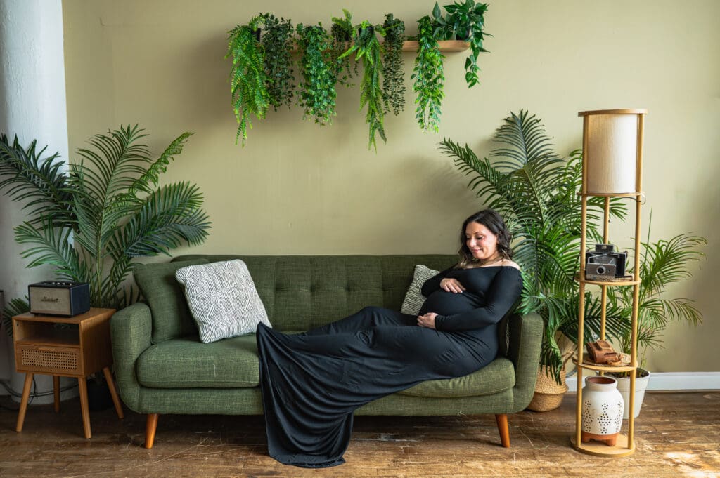 A pregnant woman in a black dress sits on a green couch holding her belly.