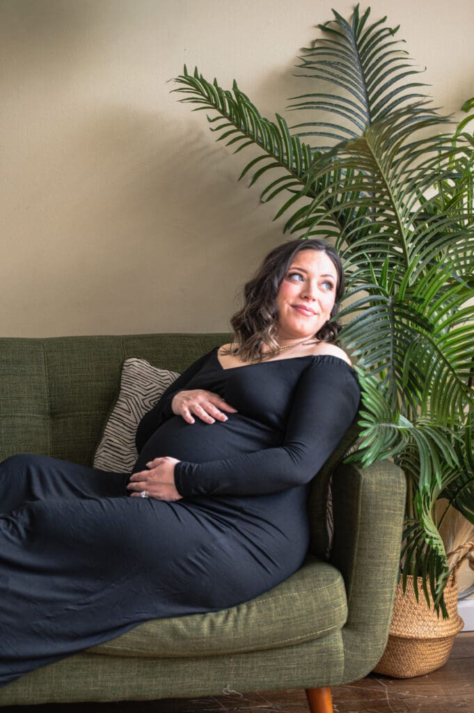 A pregnant woman holding her belly is laying on a green couch and looking back, off into the light.