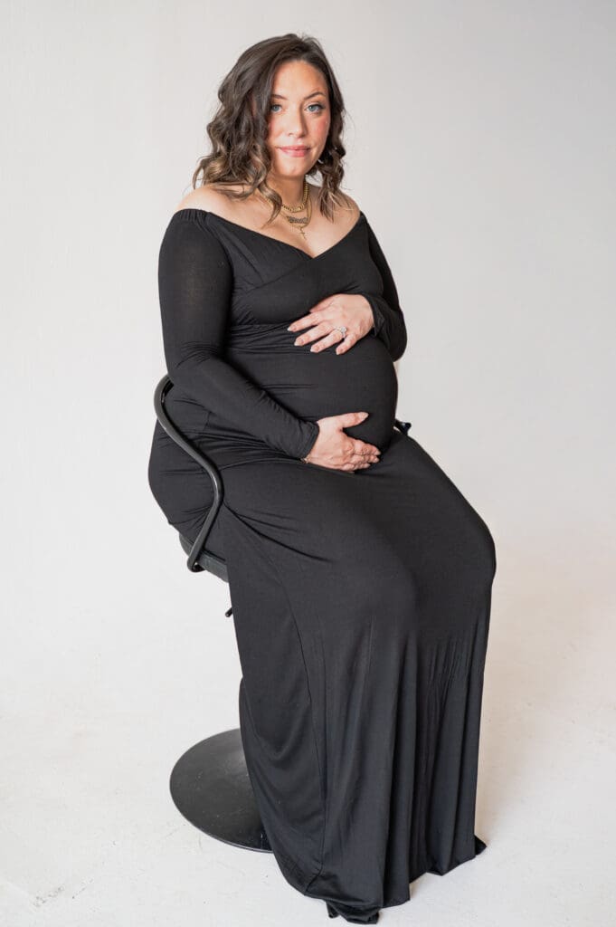 A pregnant woman in a black dress is sitting in a black chair, holding her belly, in front of a white back drop, looking directly at the camera with a straight face.