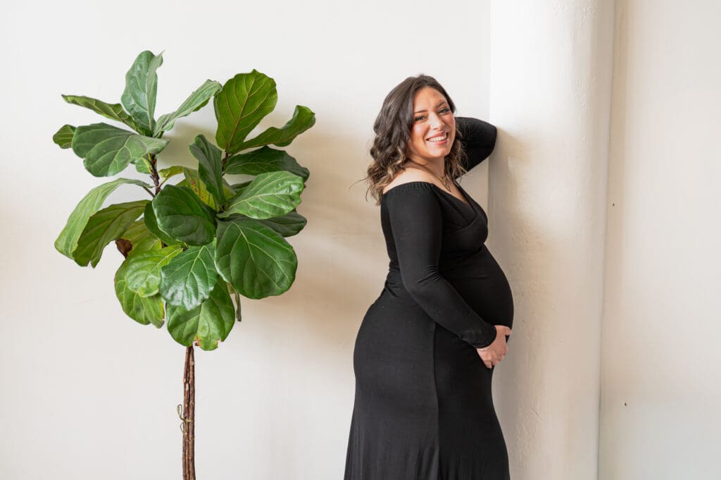 A pregnant woman leaning against a wall laughs at the camera, a green plant is behind her. 