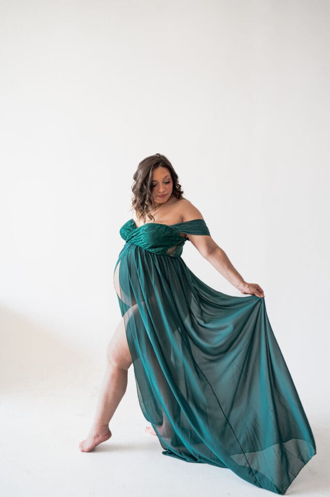 A pregnant woman in a green flowing dress is holding the tail of the dress behind her, with one leg pointed in front of her, looking back at the tail of the dress flowing. 