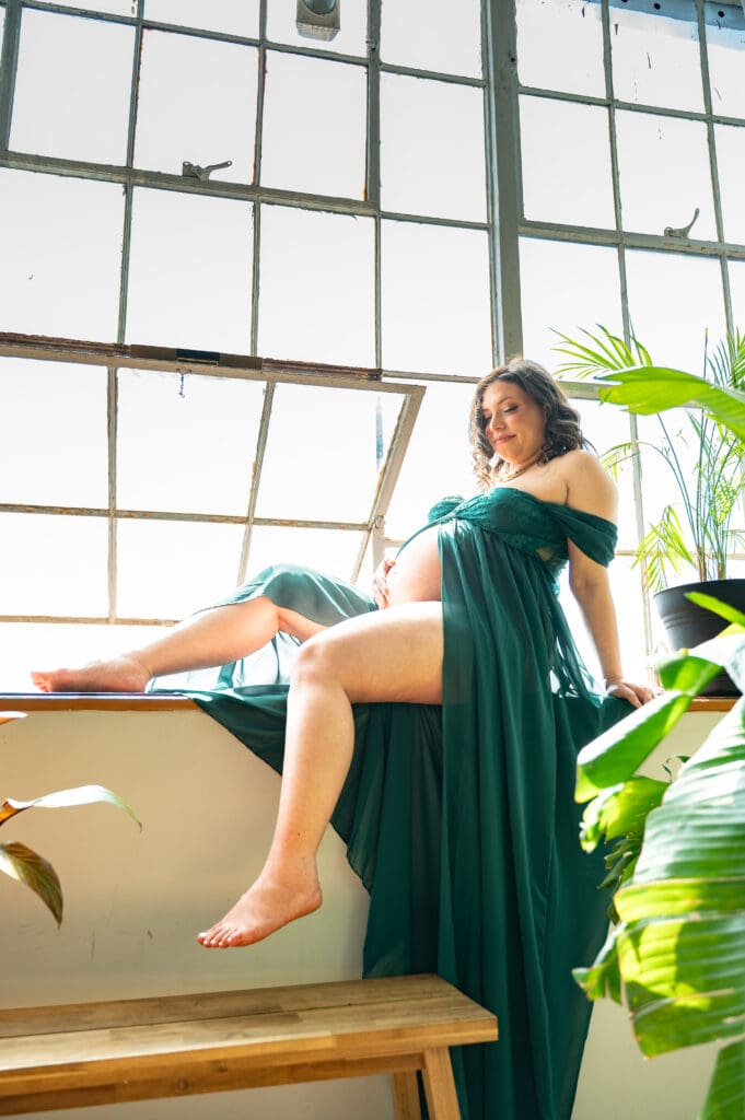 A pregnant woman in a green dress sits on the windowsill with green plants surrounding her. She is looking down at her belly.