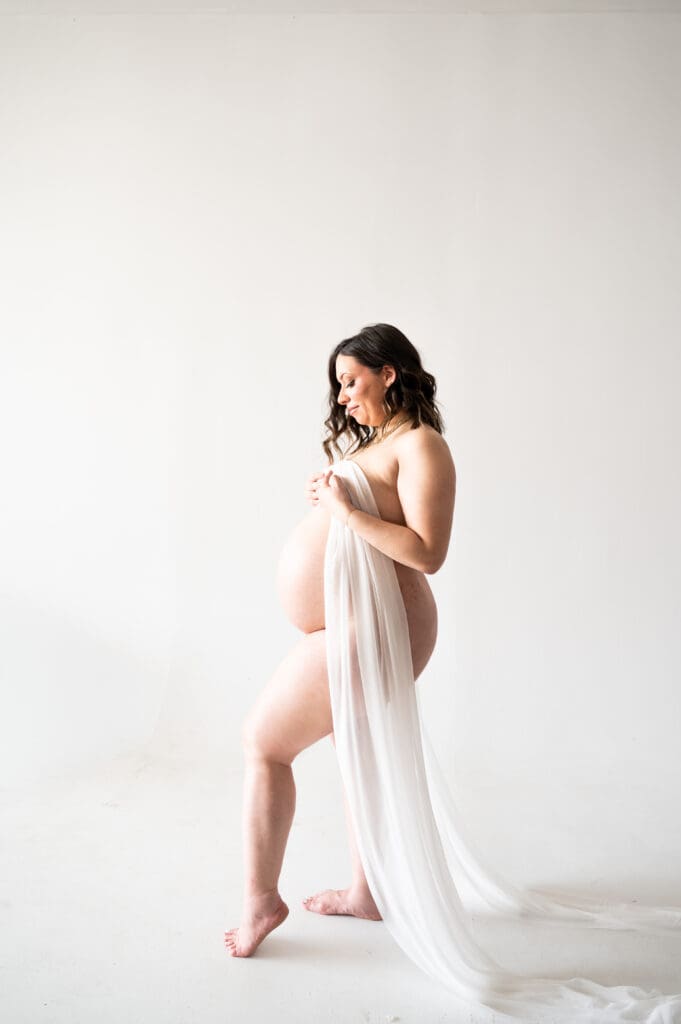 A pregnant woman standing profile to the camera, with her front leg pointed and a white sheer sash is draped over her, with her belly showing.