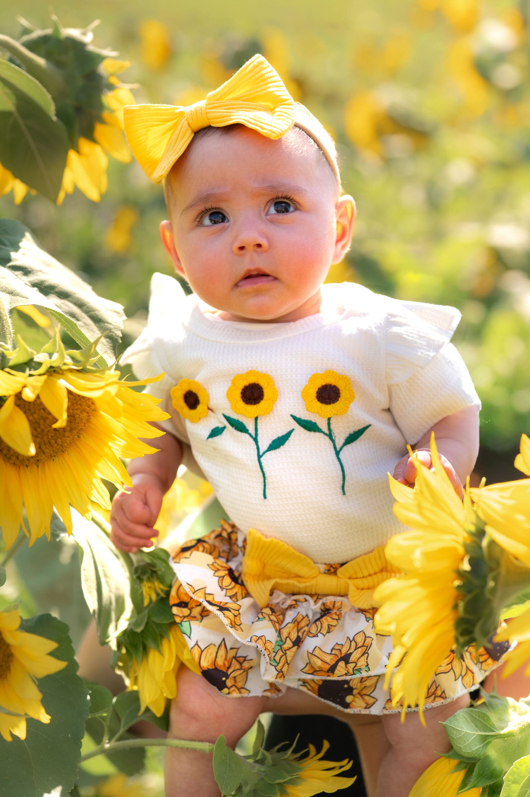 A baby wearing a sunflower outfit poses in a field of yellow sunflowers.