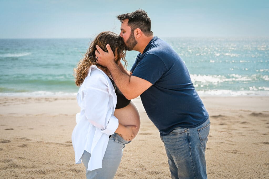 A husband kisses his pregnant wife on the forehead while posing on the beach on a sunny day for photos.