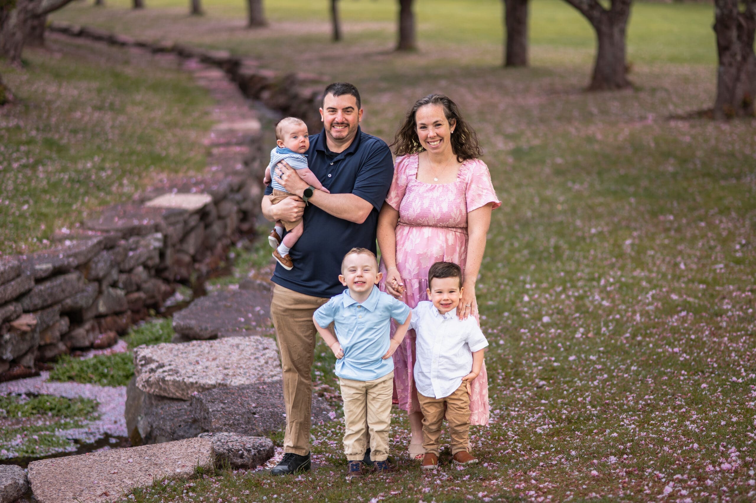 A family of five is in a park surrounded by pink leaves from cherry blossoms.
